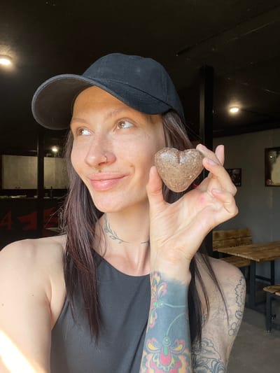 A woman with a black baseball cap, wearing a black tank top, is holding a heart-shaped rock in her right hand. She has tattoos on her left arm and her chest. She is indoors, and the background includes dark walls, lighting fixtures, metal bars, and wooden furniture. The location is Doma 45 Kilometr, Russia.