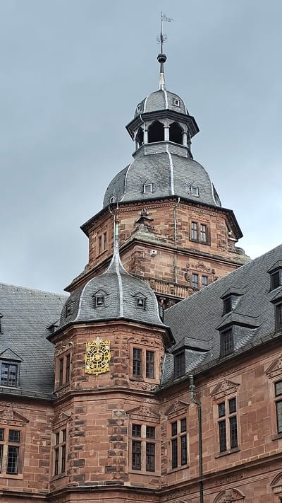 The image displays an upward, close-angled view of a section of Schloss Johannisburg (Johannisburg Palace) in Aschaffenburg, Germany, under a uniformly overcast, light grey sky. The structure is constructed from reddish-brown sandstone blocks, exhibiting precise masonry and carved details.
In the background, a prominent tower rises, capped by a large, multi-tiered dome covered in dark grey slate tiles. Above this dome, an open lantern structure with arched apertures supports a conical spire, which culminates in a finial topped by a small, banner-like weather vane. The stone sections beneath the dome feature rectangular windows with dark frames.
In the midground and foreground, a lower, intricate turret projects from the main palace structure, also clad in reddish-brown sandstone. This turret is topped by a smaller, pointed dome, similarly covered in dark grey slate, and features multiple dormer windows; one dormer clearly reveals a small bell. Centered on the turret's sandstone facade is a distinct, ornate golden clock face with a radial sunburst design, depicting Roman numerals, and the inscription "HORIS" directly below. Numerous multi-paned, dark-framed windows are set into the surrounding sandstone walls, which display rustication and decorative friezes. Portions of other steeply sloped, dark grey slate roofs with various dormers are visible in the composition. No individuals, vehicles, or other movable objects are present within the scene.