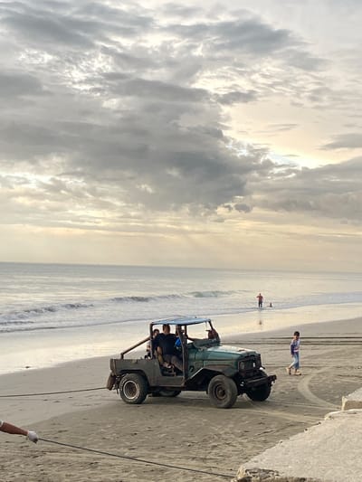 The image depicts a beach scene in Bateetimoh, Indonesia, under a cloudy sky. A vintage green jeep is positioned on the sandy shore, with several people inside. A rope is attached to the front of the jeep, and a person wearing a white glove is holding the other end, suggesting the vehicle is being pulled or is about to be moved. In the background, the ocean meets the shore with gentle waves. Further out in the water, two individuals are visible, with one standing and the other appearing to be in a crouched position. On the beach to the right of the jeep, a child walks away from the vehicle, leaving tire tracks in the sand. The overall atmosphere is overcast, with the sun faintly visible through the clouds.