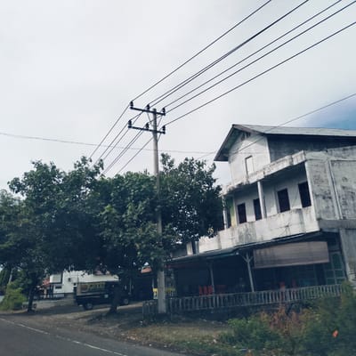 The image presents a daytime street scene in Teungoh Baro, Indonesia, under an overcast, bright sky. On the right, a two-story, light grey concrete building with a dark, corrugated metal roof stands prominently. The upper story features a single, unadorned window opening, while the lower level includes a balcony with multiple dark-framed windows and an outdoor seating area shaded by an awning. Orange chairs are visible beneath the awning, fronted by a decorative concrete railing.
A tall concrete utility pole with numerous black power lines stretches across the upper half of the frame, originating from near the center-left. Lush, dense green foliage from large trees dominates the left side and central mid-ground, partially obscuring a smaller white building further down the road. An asphalt road occupies the immediate foreground. A dark-colored pickup truck is parked on the left, partially hidden by the trees. Text visible on the side of the truck includes "MIE KOC" and "& KANJI CA". A yellow sign, partially obscured, shows text resembling "ALEU" near the utility pole. No explicit human activity is visible, depicting a tranquil, ordinary street view.