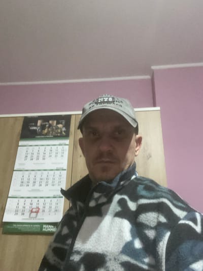This is a vertically oriented, close-up, eye-level medium shot of a man wearing a grey baseball cap and a patterned fleece jacket. The man is looking towards the camera with a neutral expression. His face is partially obscured by the angle of the shot and his clothing. He appears to be indoors, with a wall in the background that transitions from a soft pink to a lighter shade. To the right of the man, a wooden surface is visible, and on it are three calendar pages. The calendar pages are white with green text and feature grids of numbers representing days. One calendar page has an image of what appears to be a vintage vehicle on it. Visible text on the calendars includes "03.2019", "09/2019", and "MANN+HUMMEL". The lighting suggests it could be daytime, but the indoor setting and lack of windows make it difficult to determine the exact time of day or weather conditions. The overall scene appears casual and domestic.