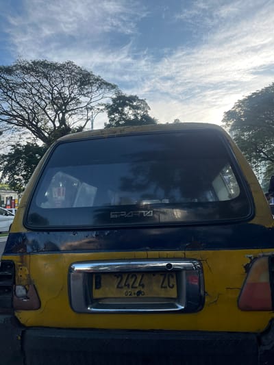 The image shows the rear of a yellow and blue truck. The truck's license plate reads "B 2424 ZC 02.26". The word "SPARTA" is visible on the rear window. The paint on the truck is peeling. The location is Kelapa Dua, Indonesia, as suggested by the context. In the background, there are trees and a partly cloudy sky. The reflection of surrounding buildings is visible on the rear window.