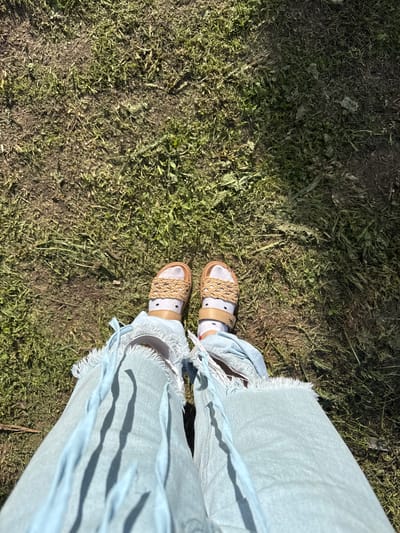This media file captures a high-angle, first-person perspective, looking down at a person's feet and lower legs. The individual is standing on an outdoor surface composed of sparse green and dried grass mixed with visible dirt and soil, indicative of a natural, possibly unkempt, ground area.
The person is wearing light blue denim pants, which feature notably frayed and tattered hems, suggesting a distressed or casual style. Their feet are clad in sandals with light brown or tan soles and woven straps made of a natural fiber, such as rope or jute, forming an intricate pattern across the top of the foot. White socks with small black polka dots are worn with the sandals.
Strong, distinct shadows are cast across the ground by the pants and sandal straps, indicating bright sunlight and suggesting the time of day is sunny, likely mid-day or afternoon. No specific activity is depicted beyond standing or being present in this outdoor setting. There is no visible text within the image. The scene conveys a casual, outdoor ambiance, fitting for a rural or semi-rural location like Луптюг, Russia.