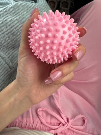 A close-up, high-angle shot shows a person's left hand gently holding a small, pink, spiky massage ball. The hand is positioned on the lower right of the frame, with the ball nestled in the palm. The person's fingernails are neatly manicured and painted a light pink. The background consists of soft, textured fabrics in shades of gray and pink. To the left, a gray, knitted fabric is visible, while to the right, a soft pink fabric with drawstring details is present. The lighting is soft and diffused, highlighting the texture of the ball and the subtle sheen on the nails. No specific location in Bashtanka, Ukraine, is discernible from the image.