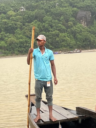The image shows a young man standing on a wooden boat in the river at Jaflong, Bangladesh. He is wearing a blue polo shirt and grey pants, and a baseball cap. He is holding a long wooden pole. The river is a light brown color and appears calm. In the background, there is a lush green hillside covered in trees. Several boats can be seen in the distance on the river. The weather appears overcast, possibly with light rain.