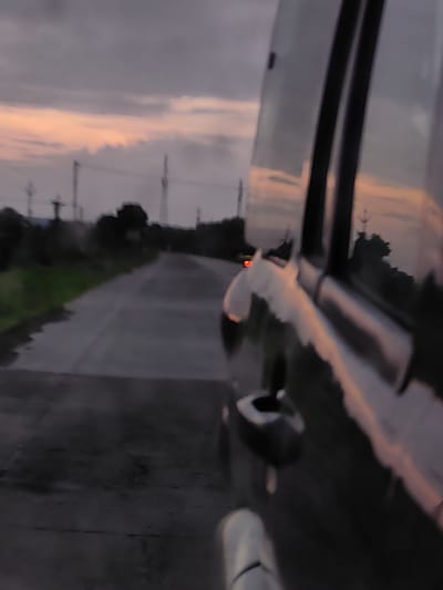 The image depicts a view from inside a vehicle, looking out at a road at dusk. The sky is a muted orange and purple, indicative of sunset or twilight, with clouds present. In the distance, there are silhouettes of trees, poles, and possibly buildings. The road surface appears wet or reflective, suggesting recent rain or damp conditions. The vehicle's interior is visible, including a door handle and possibly a window. There are no people clearly visible within the frame, and the focus is on the external environment and the reflection on the vehicle's surface. The scene appears to be on a road outside of the urban center, potentially on the outskirts of Padli, India, given the presence of rural or semi-rural elements like trees and poles.