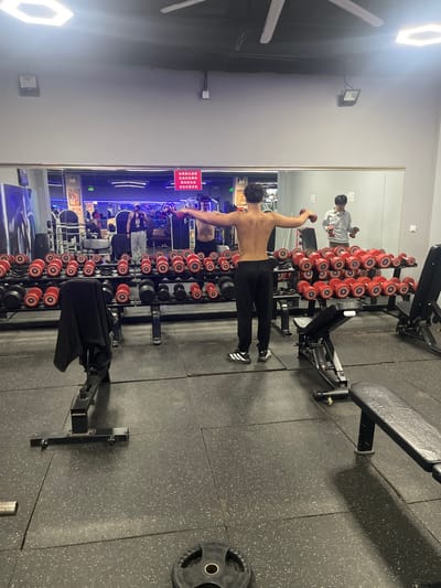 The image depicts an indoor gymnasium in Shenhe District, China. A young adult male, bare-chested and wearing black athletic pants with black and white striped shoes, stands with his back to the viewer in the foreground. He is performing a lateral raise exercise, holding a small red dumbbell in each outstretched hand, positioned horizontally.

The gym features dark grey rubberized flooring with light speckles, composed of interlocking tiles. The background consists of a large mirror wall, reflecting the gym's interior and other individuals. Directly behind the main subject, a two-tiered black metal dumbbell rack holds numerous red-ended dumbbells with silver handles, arranged in ascending/descending weight order. A second similar rack is visible to the right.

Reflected in the mirror, to the main subject's left, another male in a black hoodie and light pants appears to be near equipment. To the main subject's right in the reflection, a male wearing a light-colored long-sleeved shirt and dark pants is also holding small red dumbbells. Above the reflected dumbbell racks, a red rectangular sign with white Chinese characters is visible, likely displaying gym rules or instructions.

Overhead, hexagonal LED lights illuminate the space, along with a rectangular recessed light and a large white ceiling fan. Two black adjustable weight benches are present in the foreground; one to the left has a black garment draped over it, and another is partially visible to the right. A single black weight plate with a silver center rests on the floor in the lower left foreground. The walls are a light grey color.