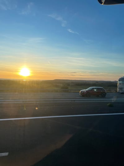 The image depicts a multi-lane highway at the approximate time of sunrise or sunset, situated near La Palme, France. A bright, intensely yellow-orange sun is positioned low on the left horizon, radiating light across the scene and causing significant lens flare. Beyond the highway, a body of water, consistent with the Étang de La Palme, is illuminated by the sun, bordered by a strip of vegetated land and distant, low-lying hills. The sky transitions from rich orange and yellow tones near the horizon to a clear light blue overhead, with sparse wispy clouds and a faint contrail visible in the upper left.
The highway occupies the lower portion of the image, appearing as dark asphalt. A solid white line marks the left edge of the immediate foreground lane. In the middle lane, a dark red compact SUV with black wheels is in motion, traveling rightward. A bright circular reflection is visible on its passenger-side window. To the right of the SUV, the front cab of a white semi-truck is partially visible, also moving rightward in an adjacent lane. Metal guardrails separate the highway lanes. No individuals are discernible. A faint shadow, likely from the vantage point of the photographer's vehicle, is cast on the foreground pavement.
