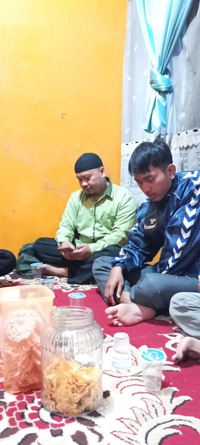 The image shows two men sitting on the floor in a room in Pameungpeuk, Indonesia, at the location -7.016, 107.567.  The man on the left is wearing a green long-sleeved shirt and black pants. He is wearing a black skullcap. He has his right hand on his lap and his left hand is holding a smartphone. The man on the right is wearing a blue jacket with white chevron stripes and grey pants.  He has a Hummer logo on his jacket. His left hand is on his lap. His right foot is bare.  He is looking down at his lap. The room has a yellow wall with a piece of furniture on the right, partially obscured. The floor is covered in a red carpet with white and black patterns. There are several glass jars and glasses on the carpet in front of the men. One jar is filled with what appears to be potato chips. Another jar is filled with what appears to be a noodle snack.  The room is brightly lit.