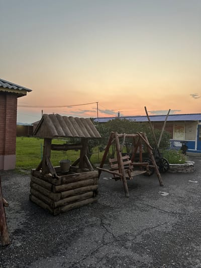This image depicts an outdoor scene at dusk, with a soft, gradient sky transitioning from pale blue at the top to shades of orange and pink near the horizon. The location is identified as Aeshka, Russia.
In the foreground and midground, a rustic wooden well stands prominently on a gravelly courtyard. The well is constructed from logs, with a tiered base and a sloped wooden roof. A metal bucket is suspended inside the well by a chain. To the right of the well is a wooden swing set, also made from rough-hewn logs, with a bench swing. Behind the swing set, a portion of a building with a blue roof is visible, adorned with what appear to be decorative panels or artwork. Further in the background, a brick building with a yellow trim is seen on the left, and a grassy field stretches towards a distant treeline and a faint outline of hills under the twilight sky. Power lines are also visible stretched across the sky. The overall impression is one of a rural or semi-rural setting, possibly a courtyard or yard at a residence or establishment. No people are present in the image.