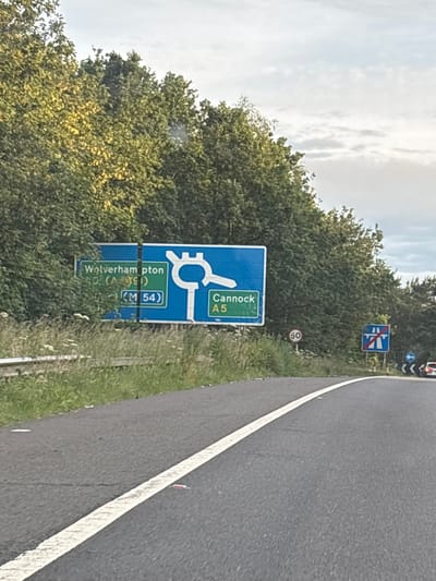 A road sign indicates directions to Wolverhampton and Cannock. The sign, which is blue and white, shows a diagram of a roundabout with arrows pointing to the destinations. "Wolverhampton" and "A5" are visible on the left side of the sign, along with "(M 54)" in a green box. On the right side, "Cannock" and "A5" are displayed. The sign is positioned next to a road in a rural or semi-rural setting, with trees and vegetation on the left and a clear sky above. A circular speed limit sign with "50" is visible below the main road sign. Further down the road, another sign indicating the end of a dual carriageway is present. A single white line marks the edge of the road. The scene is captured from the perspective of someone on the road, suggesting a driving context. The location is South Staffordshire, United Kingdom.