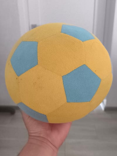 A close-up shot shows a yellow and light blue patterned foam ball being held by a person's hand. The ball has a textured surface and is partially obscuring the hand. The background is out of focus, showing what appears to be an indoor environment with white walls and some vertical lines that could be doors or curtains. The lighting suggests an indoor setting. There are no explicit geographical markers to indicate a specific city or country. The image quality is slightly grainy.