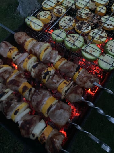 This image depicts a close-up, overhead view of a barbecue in progress. Several skewers of meat and vegetables are being grilled over glowing red coals. The meat appears to be cubed and interspersed with slices of yellow squash and possibly white onion. On a wire rack above the skewers, more sliced zucchini and yellow squash are grilling, showing char marks. The scene is set outdoors, with a hint of green grass and a dark, textured surface, likely a tree trunk or large rock, visible in the background. The lighting suggests it may be dusk or evening. The location is identified as Матыкальскі сельскі Савет, Belarus. There are no people visible in the frame.