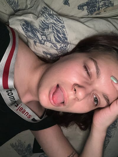 The image shows a close-up, overhead view of a young person lying down. The person has dark hair, a septum piercing, and appears to be winking with their tongue slightly out. They are wearing a black garment with a red and white striped band that says "tommy jeans". The background appears to be a bed with a patterned comforter. There are no identifiable landmarks or contextual clues that would place this image in Bishkek, Kyrgyzstan.