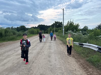 A group of people are walking and running down a dirt road in what appears to be a rural setting in Маркидоновка, Russia. The group consists of at least two adults and three children. The sky is overcast with dramatic clouds, suggesting either an approaching storm or a recent one. The trees and foliage are lush and green, indicating it is likely summer.

In the foreground, a young boy wearing a black baseball cap, a dark t-shirt with a graphic of an eagle, black pants, and pink shoes is running towards the camera. To his right, another boy in a yellow t-shirt and black pants is walking away from the camera. Further down the road, two adults are walking with two young girls. The adult on the left, a man, is wearing a dark t-shirt and black pants. The adult on the right, a woman, is wearing a blue jacket and brown pants. The two girls are dressed in casual clothing.

Other notable details include a utility pole with a sign attached on the right side of the road, and a guardrail along the side of the road. The overall mood appears to be casual, perhaps a family outing or a walk in the countryside. The time of day is likely late afternoon or early evening, given the lighting and the overcast sky. There is no visible text other than what might be on the sign on the utility pole, which is too small to read.