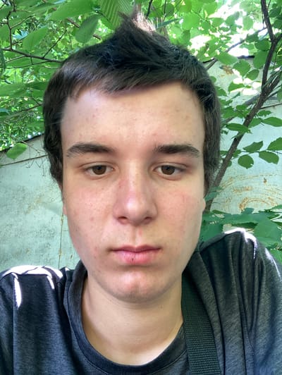 A close-up, head-on selfie of a young Caucasian male, appearing to be in his late teens or early twenties, is captured outdoors. He has brown hair styled with a spiky top and dark brown eyes. His skin shows some redness and blemishes. He is wearing a dark gray t-shirt. The background consists of lush green foliage from trees and a weathered, light-colored wall, possibly made of corrugated metal with rust spots. Sunlight filters through the leaves, creating dappled light and shadows on his face and clothing. The scene appears to be in a natural, possibly overgrown, urban setting. Based on the available information, the photo is geotagged to Pervomaisk, Ukraine.