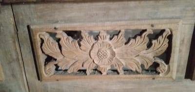 A wooden decorative panel featuring a floral motif is affixed to a larger wooden surface. The panel is rectangular and has an openwork design with a central flower surrounded by stylized leaves. The wood appears unfinished and slightly weathered. The panel is attached to the larger surface with nails visible in the corners. The background is dimly lit, suggesting an indoor setting. While the exact location cannot be determined solely from the image, the prompt places it within the city of Bulukandang, Indonesia.