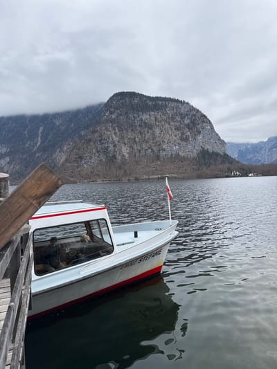 A white motorboat, identified as "STEFANIE" with registration "O-10322," is docked at a wooden pier on Hallstätter See in Hallstatt, Austria. The boat features a white hull with a red stripe at the waterline, a grey deck, and an enclosed cabin. An Austrian flag (red-white-red) is affixed to a pole at the boat's bow. Two individuals are visible within the cabin through a large window: one person in dark clothing is seated on the left, and another individual with blonde hair is partially visible to the right. The boat is positioned in the lower-left of the frame, partially obscured by a diagonal wooden beam of the pier. The lake's surface is rippled, reflecting the boat and the cloudy sky. In the background, steep, forested mountains with visible rock faces and sparse winter vegetation rise directly from the lake. Patches of snow are discernible on higher elevations and in shaded areas of the mountains. The upper sections of the mountains are partially enveloped by low-hanging grey clouds, consistent with the overcast sky. Several small structures are visible on the distant shoreline to the right.