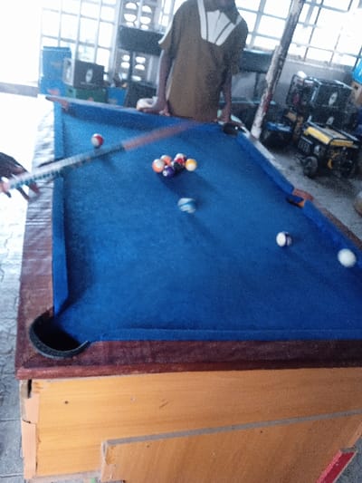 The image depicts an indoor scene in Barama, Nigeria, where two individuals are engaged in a game of pool. The central element is a blue-felt pool table, featuring a dark brown wooden frame and lighter wooden legs that show signs of wear.
On the left side of the frame, a person's hands are visible, holding a pool cue stick which appears blurred due to motion, indicating a shot is being taken. The cue ball, striped blue and white, is seen in the path of the cue. A cluster of racked pool balls, including solids and stripes of various colors, is positioned further down the table. Other individual balls, including a red and white striped ball and a solid white ball, are scattered across the blue felt.
Another individual stands on the opposite side of the pool table, partially visible from the chest down. This person is wearing a brown shirt with a distinct white, wing-like pattern on the collar and appears to be observing the game, with their hands resting on the table's edge.
The background reveals a functional, perhaps utilitarian, indoor space. Bright natural light streams in from large windows at the top of the frame. Various items are stored in the room, including several portable generators of different models and sizes, along with what appear to be black speakers or storage containers. The general ambiance suggests a casual, local gathering spot or a community game room. No discernible text is visible on any of the objects or surfaces. The time of day appears to be daytime, given the natural light.