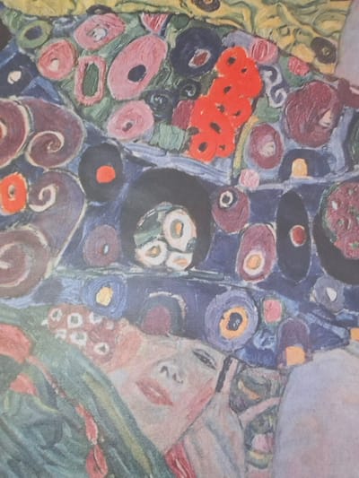 This image is a close-up of a painting, likely a detail from a larger work, featuring a stylized, semi-abstract depiction of a woman's face partially obscured by ornate, patterned fabric. The painting uses a rich palette of colors including pinks, reds, blues, purples, greens, and yellows. The style is reminiscent of Art Nouveau or Symbolism, with swirling patterns and organic shapes.

The visible part of the woman's face shows a closed eye, a stylized nose, and full lips painted in a soft pinkish-red hue. Her skin tone is rendered in pale peaches and pinks. She appears to be wearing elaborate clothing or adornments that are part of the overall composition.

The background and surrounding elements are a complex arrangement of circular and floral motifs, as well as swirling, scroll-like patterns. These elements are rendered in bold colors and textures, creating a dense and decorative surface. Some of the circular motifs contain darker centers, resembling eyes or abstract floral centers. There is a cluster of bright red, blob-like shapes in the upper right quadrant of the image.

The painting is exhibited at Serlachius Museums in Mänttä-Vilppula, Finland.