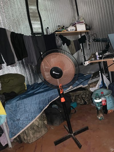 The image depicts an indoor, cluttered scene with various objects arranged in what appears to be a temporary living space or storage area. The setting seems to be a room with walls covered in a textured, possibly insulated material, giving it a somewhat utilitarian or unfinished feel.

Dominating the foreground is a large, circular fan with a brown-toned grille and a black base. Attached to the fan's stand is a black pole with an orange tip, possibly an extension or part of a different device. Scattered around the fan and extending into the background are piles of clothing, including what looks like dark-colored pants and shirts, as well as some items in camouflage patterns. A rolled-up sleeping bag, olive green in color, is also visible.

On a shelf to the left, several items are placed, including a white box and what appears to be a small white appliance with three dots. Above this shelf, a computer monitor is mounted on an adjustable arm.

In the mid-ground, several water bottles are visible, one red-labeled Coca-Cola bottle and a clear bottle with a green cap. These are placed among bags and fabric items. A large blue quilted blanket or sleeping bag covers a significant portion of the right side of the image. Further back, a piece of metal equipment with a cross-shaped base stands upright.

The lighting suggests it might be nighttime or a dimly lit interior space, as there are no strong natural light sources. The overall impression is one of disarray and a lack of organization, indicative of a space being lived in or used for storage under less than ideal conditions. The presence of sleeping bags and clothing suggests it's a place for rest or shelter.

Visible text includes "BUILT" on the Coca-Cola bottle label and "DOMOTEC" on the fan's motor housing. There is no indication of time of day, weather, or specific emotions. The location context provided is Martove, Ukraine.