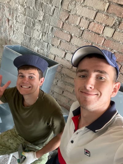 Two young Caucasian males are taking a selfie in what appears to be a construction or renovation site. The background consists of a rough brick wall and large metal ducts. The male on the left is wearing a dark green t-shirt, camouflage pants, a blue flat cap, and white gloves. He is smiling broadly and holding up two fingers in a peace sign. He is holding a trowel in his gloved left hand. The male on the right is wearing a white polo shirt with a navy collar and red accents, and a blue baseball cap. He is also smiling. The overall impression is that of a break during manual labor. The location is identified as Аснежыцкі сельскі Савет, Belarus.