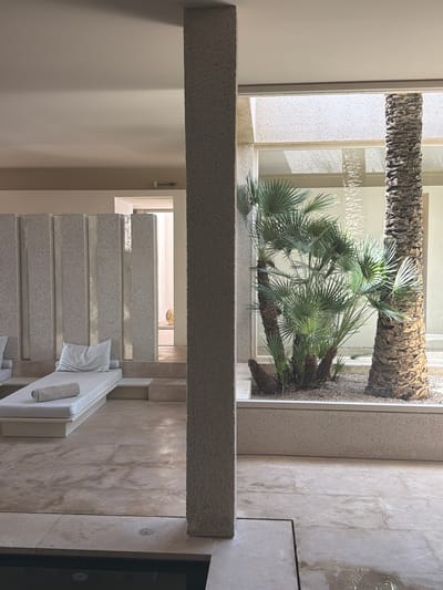 The image depicts an indoor spa or relaxation area with a minimalist design. A large, textured concrete pillar dominates the foreground, bisecting the scene. To the left, a white lounge chair with a rolled towel and a white pillow is placed on a raised platform, against a wall of vertical concrete elements. Beyond this, a doorway leads to a hallway. To the right of the pillar, a large window looks out onto an indoor courtyard with two palm trees and lush green foliage. The floor is made of light-colored stone tiles. The overall atmosphere is serene and modern. The specific location is not discernible from the image itself, and no people or actions are visible.