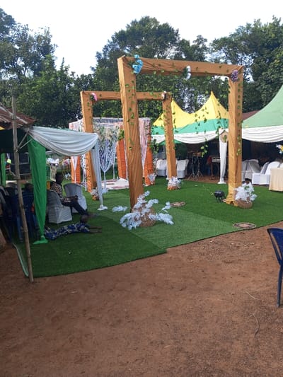 In Amokvo, Nigeria, an outdoor event setup is observed under daylight conditions. The scene is bisected horizontally; the foreground consists of reddish-brown bare earth, while the mid-ground features a rectangular area covered with green artificial turf.
On the artificial turf, a series of four large, square-columned archways are erected. Each column is wrapped in light brown fabric, decorated with green artificial vines, and topped with clusters of artificial flowers in blue, pink, and purple hues. Between some archways, white draped fabrics and vertical orange banners are visible.
To the left, a rudimentary tent structure stands, composed of bamboo poles supporting draped green and white fabric. Beneath this structure, on the artificial turf, one individual is lying prone, facing away from the camera, wearing a patterned blue and gold long-sleeved garment with arms extended forward. Directly behind this individual, another person sits on a white plastic event chair, clad in patterned attire. Further to the left, partially obscured, are additional individuals and a stack of white plastic chairs.
Beyond the archways, multiple event tents are present. One features yellow and green striped fabric with white draping. Another, partially visible, appears green with white draping. Under these tents, white plastic chairs are arranged, some around tables covered with light-colored cloths. A musical instrument setup, including a drum kit, keyboard, and speaker, is visible under one tent. On the artificial turf, several white artificial plant decorations, resembling branching structures, are placed, some within woven basket-like bases. A black stage lighting fixture is also positioned on the turf. The background consists of dense green foliage from mature trees under a bright, overcast sky.