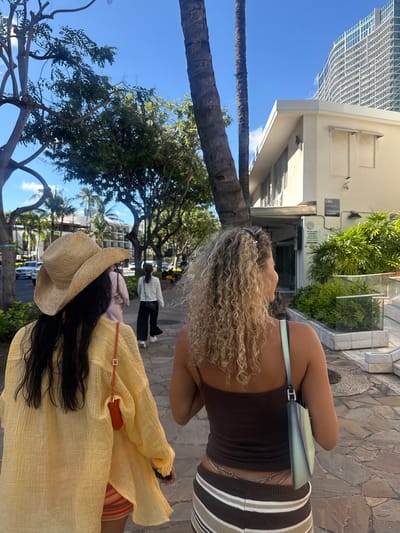 On a bright day in Honolulu, United States of America, a street scene captures two women walking. One woman has blonde, curly hair, a brown top, and a striped skirt, carrying a pale green purse. The second woman wears a straw hat and a long, yellow shirt, with dark hair cascading down her back. The background features a mix of urban and natural elements: a tall building, palm trees, and various other trees line the street. Other pedestrians are visible in the distance.