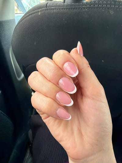 The image displays a close-up, angled view of a person's light-toned left hand. All five visible fingernails are manicured in a classic French style, featuring a glossy, translucent pink or nude base coat and precisely applied opaque white tips, primarily in an oval or almond shape. The hand is held still, without visible jewelry or specific action. The background is indistinct, composed primarily of a dark, textured fabric, consistent with the upholstery of a car seat, including visible stitching. A lighter, blurry area in the upper left background suggests ambient light from an exterior environment. The image contains no visual elements to confirm the specified location as the city of Galil Elyon Regional Council, Israel.