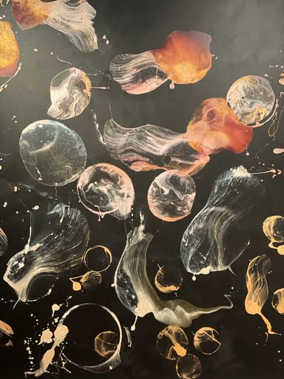 This is a vibrant, abstract close-up image of a fluid art painting. The dominant background is a glossy, deep black, providing a strong contrast to the colorful, organic shapes and patterns. The artwork features an array of swirling, dripped, and splashed forms in metallic gold, copper, white, and pale pink, with some hints of silver or light blue.
The shapes vary from distinct circles and rings, some appearing like bubbles or cells, to more amorphous, flowing designs that suggest movement and fluidity. Within these shapes, the colors are often swirled together, creating intricate marble-like textures and iridescent effects, particularly with the metallic paints which catch the light. Numerous small splatters and drips of paint, primarily white and pink, are scattered across the black surface, adding to the dynamic composition.
The overall impression is one of shimmering liquids in motion. The glossy finish suggests a resin coating or a similar technique that enhances the depth and sheen of the colors. There are no people, recognizable objects beyond the paint itself, or visible text in the image. No specific time of day, weather, or discernible location cues are present in the artwork itself.