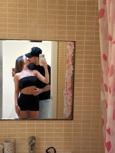 A selfie is captured in a bathroom mirror reflecting a couple kissing. The woman has long brown hair and is wearing a black tube top and a black mini skirt. The man is wearing a black baseball cap, and a black t-shirt. He is holding up a cell phone. The bathroom has cream-colored square tiles and a pink and white shower curtain. A hair tie, faucet, and decorated container are visible on the right side of the frame. It is not possible to establish the specific location in Platja D'alcúdia, Spain, based on the image alone.