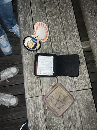 A wooden picnic table holds a variety of objects. A scallop shell with a round object inside rests near an open box with a black felt lining and paperwork. A square metal tin with a faded design of crossed sticks within a circle lies nearby. Parts of three pairs of feet in sneakers are visible at the bottom of the frame, indicating the perspective is from above. The scene is generally dimly lit.