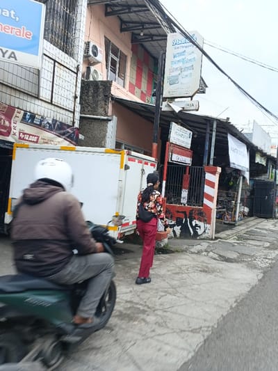 A street scene features a motorcyclist with a white helmet and grey attire, alongside a person in a floral shirt and red pants. Buildings line the street, adorned with signs in a foreign language, like "Radi" and "KRETEK." Graffiti art marks a wall near a gate. A white truck with a box body is parked nearby. The overall view suggests a busy urban environment.