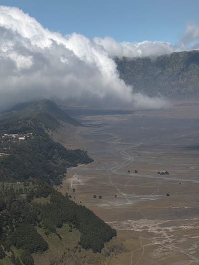A high-angle, wide shot captures a vast, arid plain stretching towards a range of mountains, with a thick band of white clouds obscuring the tops of the peaks and the sky above. The landscape is predominantly brown and grey, with scattered patches of dry vegetation and a winding dirt road leading into the distance.
To the left of the image, a steep, tree-covered hillside slopes down towards the plain. At the crest of this hill, a cluster of buildings, possibly a village or resort, is visible, nestled among the trees. The trees are dense and dark green, creating a stark contrast with the barrenness of the plain. The hillside itself is uneven, with shadowed ravines and lighter-colored patches of grass or scrub.
In the mid-ground, the plain is textured with subtle patterns of erosion and dry riverbeds. A few isolated trees dot the landscape, their dark forms standing out against the pale earth. The winding dirt road is a prominent feature, snaking across the plain and leading towards the mountains.
The clouds are a dominant element in the upper portion of the image, their thick, billowy forms creating a dramatic contrast with the clear blue sky that peeks through in some areas. The clouds appear to be hanging low over the mountain range, suggesting a misty or foggy atmosphere in that region. The overall impression is one of a rugged and remote natural landscape, potentially a volcanic caldera or a similar geological formation. The presence of the buildings suggests human habitation or tourism in this otherwise wild environment, likely near the village of Cemorolawang in Indonesia, known for its proximity to the Bromo Tengger Semeru National Park.