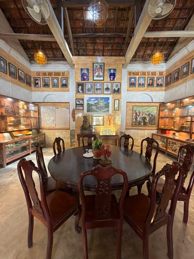The image shows an interior scene of what appears to be a museum or historical exhibit room in Hội Tín, Vietnam. A large, dark, oval wooden table with eight ornately carved wooden chairs surrounds it. On the table, there are a few objects, including a small plant and two glasses.

The walls of the room are lined with framed photographs and paintings. On the left wall, there are display cases filled with artifacts, including statues and what appear to be religious items. The ceiling is made of wooden beams and terracotta tiles, with two ceiling fans and woven bamboo light fixtures.

On the back wall, there are multiple rows of framed portraits, many of which appear to be of religious figures. There are also two large paintings on the walls, one depicting a group of people and another of a building. In the center of the back wall, there is a large framed display with a grid of small images.

The overall impression is of a well-preserved historical space, likely dedicated to showcasing the history and figures associated with the Hội Tín area. The lighting is warm, contributing to a sense of history and reverence.