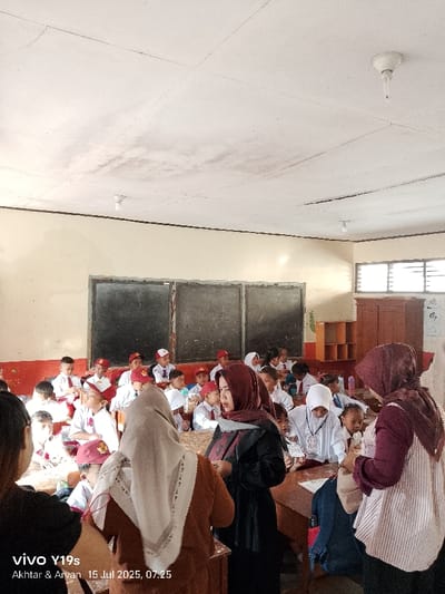 The image depicts an indoor classroom located in Jagapura Kulon, Indonesia, captured on July 15, 2025, at 07:25. The scene contains approximately 25-30 individuals.
**People**:
* **Children**: The majority are primary school-aged children, identifiable by their uniform of white short-sleeved shirts, red ties, and red caps or headwear. They are seated at wooden desks and benches arranged in rows.
* **Adults**: Three adult women are prominent in the foreground and midground. All wear hijabs. One woman, positioned centrally, is seen from behind, wearing a light brown top and a light-colored, patterned hijab. To her right, another woman in a dark top and maroon hijab is partially turned towards the left, interacting near a desk. On the far right, a third woman wearing a white and maroon striped top and a maroon hijab is also positioned near desks, seemingly engaged with the area.
**Environment and Objects**:
* **Walls**: The classroom walls are two-toned, featuring a lower red band and an upper cream-colored section.
* **Ceiling**: The white panel ceiling displays multiple irregular dark stains, indicative of water damage or discoloration, with visible seams between panels. Two basic white light fixtures are mounted, one containing a coiled fluorescent bulb.
* **Windows**: A series of horizontal louvered windows are situated on the right-side wall, providing natural light.
* **Blackboards**: Two large, dark rectangular blackboards are mounted on the rear wall.
* **Furniture**: Wooden desks and benches are systematically arranged. A multi-compartment wooden cabinet is visible in the rear right corner of the room.
**Actions and Interactions**:
The adult women are standing among the seated students, appearing to supervise or engage in instruction. The students are primarily seated, observing the adults or their immediate surroundings, indicating an active classroom session.