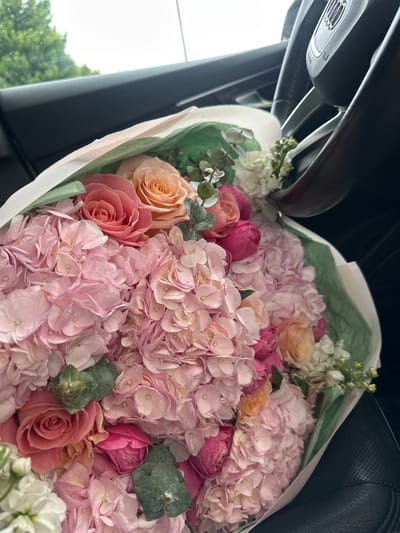 A bouquet of pink hydrangeas, roses, and white flowers is arranged in a car. The flowers are wrapped in green and white paper. The steering wheel of the car is visible in the background. The car appears to be parked on a street in Hoffman Estates, United States of America, with trees and foliage visible through the car window.