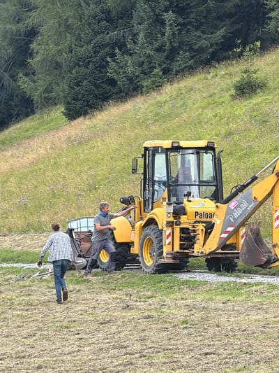 Two individuals are in a grassy field with a yellow backhoe loader. The person in the foreground, walking away from the camera, wears a blue and white checkered shirt, blue jeans, and dark shoes. The second person, closer to the backhoe, is a male with graying hair, wearing a gray t-shirt, blue pants, and dark boots. He is standing next to the backhoe, with his right hand extended towards its arm. The backhoe has the brand name "Paloar" visible on its arm. Behind them, a sloped, green hillside is covered with grass and small purple flowers, with a dense line of dark green coniferous trees at the top. The ground in the foreground is mostly dry, mown grass. The scene appears to be a rural or mountainous area in Lateis / Latais, Italy.