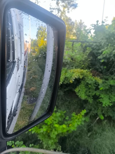 This is a close-up, eye-level, vertical shot of a black side-view mirror on a vehicle, reflecting the surrounding environment. The mirror's surface is covered with numerous small water droplets. In the reflection, the side of a white vehicle is visible on the left, with vertical lines suggesting paneling. The background reflection shows a bright sky, trees with green and yellow foliage, and what appears to be a thin vertical pole or structure. To the right of the mirror, outside the reflection, is a dense area of green foliage, likely bushes or trees, with some lighter green leaves interspersed. The scene suggests an outdoor setting, possibly after rain, in an urban or suburban environment, but no specific landmarks of Hoogstraten, Belgium are identifiable from this image.