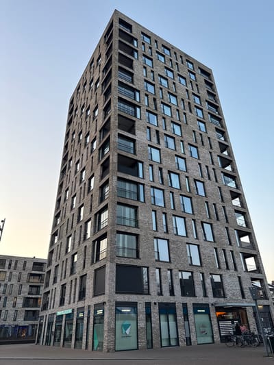 The image displays a tall, multi-story, modern brick building identified as part of the Hoge Mosten complex at Hoge Mosten 2 in Breda, Netherlands. The building features a dark grey-brown brick facade with an irregular arrangement of varying-sized windows, reflecting a pale blue and yellow sky, suggesting late afternoon or early morning conditions. Numerous recessed balconies with dark railings are integrated into the upper floors. The ground level accommodates several commercial establishments, including "Mondzorgcentrum Breda" (a dental clinic visible with "mondzorgbreda.nl" and a green tooth-like logo), a "Kunstgebit" (dentures) clinic, and a partially visible fitness center. A dark grey paved area fronts the structure. Several bicycles are parked on the right side of the frame. One individual is partially visible standing near the parked bicycles, and another person is faintly discernible inside a ground-level business. A shorter, similarly constructed brick building is visible in the background to the left.