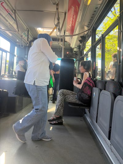 This image depicts the interior of a public bus on a bright, sunny day, with lush green trees visible through the windows, suggesting a spring or summer setting. The bus operates in Longueuil, Canada, as indicated by the French text displayed throughout the cabin.

In the foreground, a person stands with their back mostly to the camera, wearing a white long-sleeved shirt, blue jeans, and a light blue head covering. They are holding onto a vertical silver pole for stability and wearing grey athletic shoes.

Further into the bus, a young individual is seated in an aisle seat. They have dark hair, are wearing headphones, a dark top with a graphic design, and distinctive leopard print leggings. They are looking down at a can or container held in their hands, with a dark backpack visible on their shoulders. Black sandals are on their feet.

Other passengers are discernible further back, some seated and some standing, though their faces are largely obscured. One person in a black hat is partially visible seated, and another in a black shirt and green garment is standing. The bus interior features grey seats with patterned upholstery, silver handrails, and an overhead ventilation system. Sunlight streams in, illuminating the grey floor.

Visible French text includes a prominent red banner near the ceiling that reads, "Pour une virée à Longueuil en un clin d'œil" (For a trip to Longueuil in the blink of an eye). Other signs include "BIENVENUE À BORD!" (Welcome Aboard!) and a notice stating, "NOUS VOUS INFORMONS QU'IL EST INTERDIT DE Faire preuve de violence" (We inform you that it is forbidden to show violence). The scene captures a typical, calm commute on public transit.