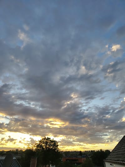 The image depicts a cloudy sky over the city of Nederbrakel, Belgium, likely during sunset or sunrise. The upper portion of the sky is dominated by large, grey clouds, interspersed with patches of lighter blue sky. Towards the horizon, the clouds are illuminated by warm, golden light, suggesting the sun is low in the sky.
In the foreground, the roofs of buildings and a few trees are visible. The roof on the far right appears to be tiled, with a dormer window. To the left, other building structures and greenery are present. A large, silhouetted tree stands prominently in the mid-ground, its branches etched against the bright sky. The overall scene captures a natural, atmospheric moment in the Belgian landscape. There are no people or animals visible in the image, and no discernible actions or interactions are taking place. The environment is that of a developed area, likely residential or a small town, given the presence of buildings and infrastructure.