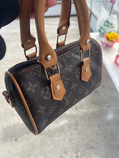 A close-up image depicts a small, barrel-shaped handbag held by a person's hand in Vlado Trichkov, Bulgaria. The handbag features a dark brown monogram pattern, characteristic of Louis Vuitton designs, with tan leather-like trim, piping, and handles. Silver-colored rivets and D-rings secure the handles to the bag's body. A dark zipper closure is visible along the top. The background, out of focus, shows a light gray ground surface. To the right, a white plastic bag with green lettering is visible, alongside arrangements of yellow and purple flowers on a white surface, consistent with a retail or market environment.