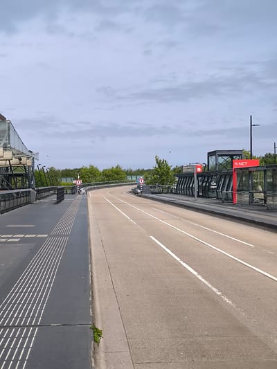 This is a view of the R-NET bus station at Hoofddorp train station in the Netherlands. The photo is taken at a high angle, on an elevated walkway, looking over a small road towards the bus station.

The bus station is on the right, marked with a large red "R-NET" sign. It has modern shelters with glass and steel construction. People are waiting inside the shelters.

The road has white lane markings and is empty of vehicles at the time the photo was taken. A pedestrian walkway with tactile paving is on the left. The sky is overcast. Green foliage is visible in the distance. There is a pedestrian crossing sign visible on the opposite side of the road.