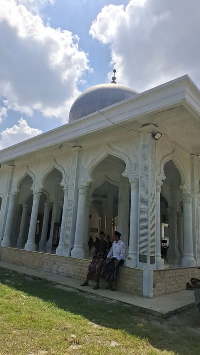 Two men are sitting on a low stone wall in front of a white mosque in Grong-grong, Indonesia. The man on the left is wearing a dark brown patterned sarong and a dark shirt. The man on the right is wearing a black sarong and a white shirt with black embroidered patterns and a black cap. The mosque has a dome and is decorated with intricate carvings around its arches. The sky is blue with scattered white clouds. The ground in front of the mosque is covered with green grass.