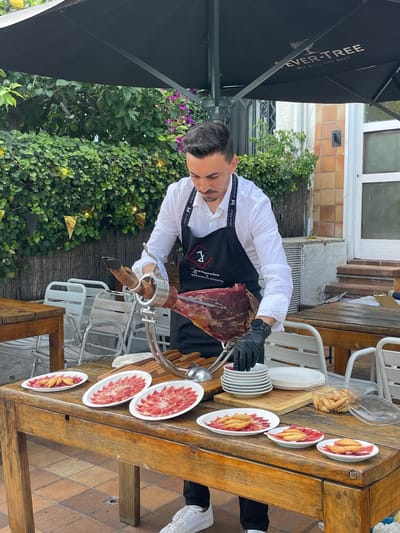 A man, wearing a white shirt, black apron, and black gloves, is slicing a large piece of cured ham at a wooden table outdoors. He is positioned under a large black umbrella with the text "EVER-TREE MIX WITH THE BEST" printed on it. The table is laden with plates of sliced ham, some topped with small, toasted breadsticks. In the background, there are white chairs, lush green foliage, and a brick building. The man is actively engaged in the task of carving the ham, holding a knife and the ham itself. The scene appears to be set in a restaurant or event space, possibly in Arenys de Mar, Spain, given the context.
