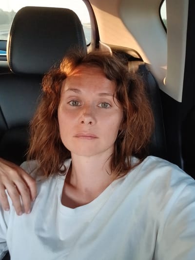 A woman with reddish-brown, wavy hair is pictured from the chest up, seated in the back seat of a vehicle. She is wearing a white t-shirt. Her left arm is bent, with her hand resting on her chest. The background shows the interior of the car, including a dark headrest and a white pillar, with sunlight casting a warm glow across the scene. The image appears to be a selfie. The metadata of the image indicates that it was taken in Teplyaki, Russia.