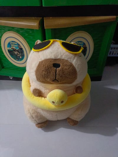 This is a photograph of a stuffed toy, likely a dog or a bear, sitting on a light-colored, tiled surface. The toy is predominantly light brown and cream-colored, with a darker brown circular patch on its face where the nose and mouth are depicted. It is wearing large, bright yellow sunglasses with black lenses and has a yellow, ring-shaped accessory around its neck, from which a smaller yellow, duck-like toy hangs. The toy's paws are visible, resting on its lap.

Behind the toy are two large, green plastic containers, possibly storage bins or tubs. The one on the left has a circular label with a blue border and white background, featuring an image of sheep and text that reads "Shaun the Sheep". The container on the right also has a similar circular label, though the details are less clear. The top edge of these containers is a darker, black color, and above that, a beige or light brown border is visible.

The lighting suggests an indoor setting, possibly with natural light contributing to the overall illumination. The shadows cast by the toy and the background elements indicate a light source from the front or slightly to the side. There are no clear indicators of time of day or weather conditions, as the scene is indoors. The presence of the "Shaun the Sheep" branding on the labels suggests a connection to the popular animated children's show. The overall mood conveyed by the image is playful and perhaps a bit whimsical, due to the toy's attire and accessories. The setting appears to be a domestic or storage area in Meunasah Raya, Indonesia.