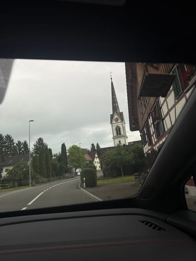 The image, captured from inside a vehicle, shows a scenic view of a village in Hugelshofen, Switzerland. The scene is dominated by traditional Swiss architecture, including a half-timbered building with white plaster and dark wooden beams, and a prominent church steeple with a clock face. The church's white tower contrasts with its dark, pointed roof, and it stands against a cloudy, overcast sky, suggesting either early morning or late afternoon, or simply a day with heavy cloud cover.

The image also features a winding road, typical of rural Swiss landscapes, bordered by lush green trees and manicured hedges. A streetlamp is visible along the road, indicating it's an inhabited area. The overall atmosphere is serene and picturesque, characteristic of the Swiss countryside. There are no people visible in the frame, and no text is present. The quality of the image suggests it was taken with a smartphone.